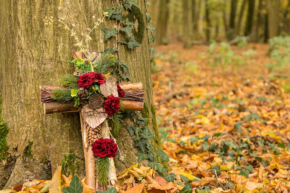 geschmücktes Holzkreuz an einem Baumstamm auf einem Waldfriedhof / Friedwald